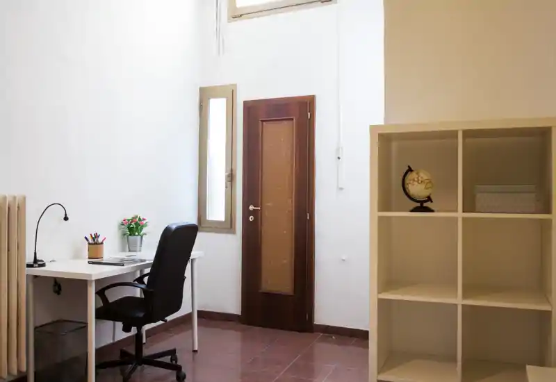 Piccolo e ordinato angolo lavoro con scrivania bianca, sedia girevole e libreria a cubi; luce naturale da una finestra in alto illumina l'ambiente.