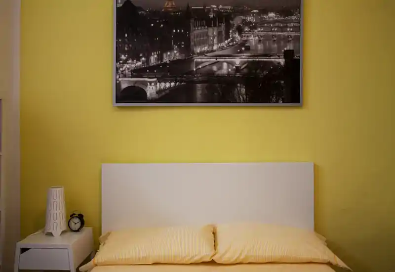Camera da letto luminosa e accogliente con testiera bianca semplice, biancheria gialla e una foto in bianco e nero della città appesa sopra il letto — perfetta per un annuncio di stanza singola o doppia.