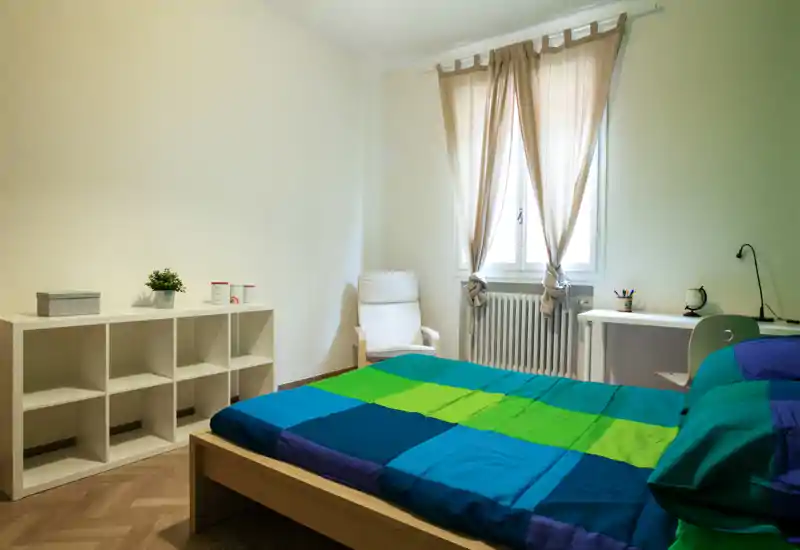 Dormitorio luminoso y de mobiliario sencillo con cama doble de ropa azul-verde, estantería blanca, escritorio junto a la ventana y sillón cómodo.
