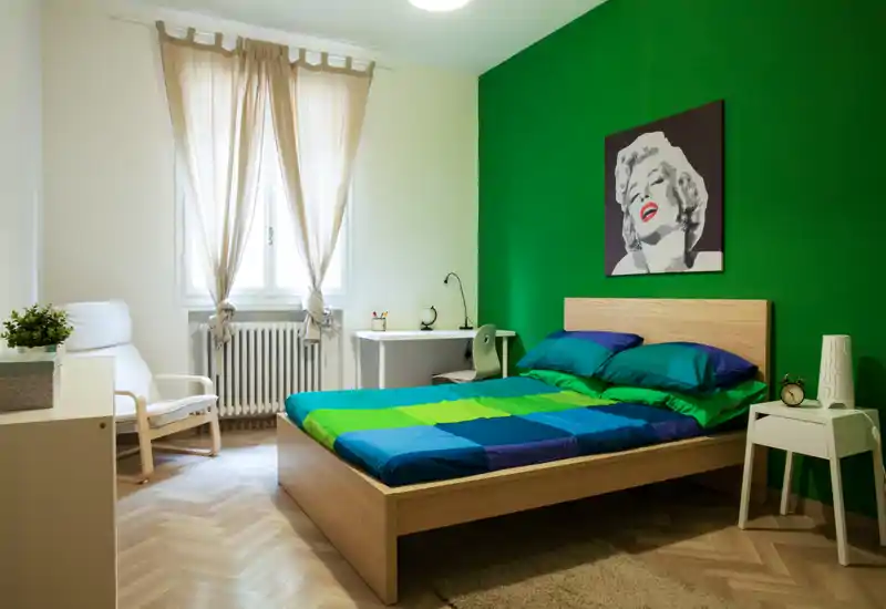Dormitorio moderno y luminoso con cama de madera, ropa de cama colorida y una llamativa pared verde; incluye un escritorio junto a la ventana y un sillón cómodo.
