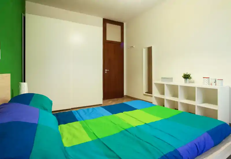Dormitorio luminoso y bien compuesto con una gran cama doble de ropa de cama colorida, un armario blanco y estantería abierta para almacenaje — ideal para un alquiler acogedor.