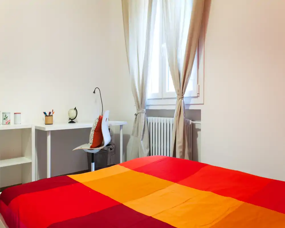 Camera da letto luminosa e accogliente con copriletto rosso e arancione, piccolo scrittoio e sedia accanto alla finestra con tende leggere — ideale per rilassarsi o lavorare.