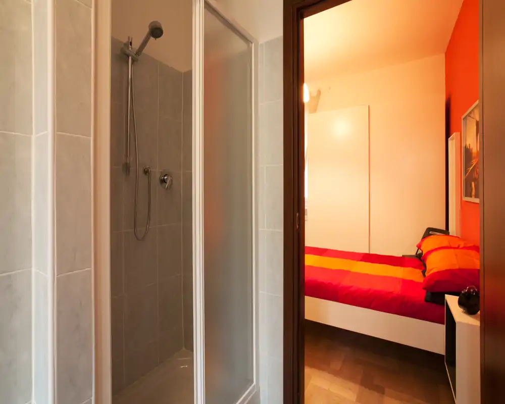 Dormitorio compacto visto a través de una puerta abierta con edredón rojo y naranja y luz cálida; mobiliario moderno y mesita de noche que hacen el espacio acogedor.