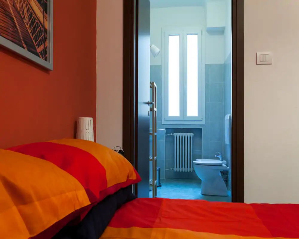 Rincón de dormitorio luminoso y acogedor con una cama colorida en primer plano y baño en suite visible a través de la puerta abierta.