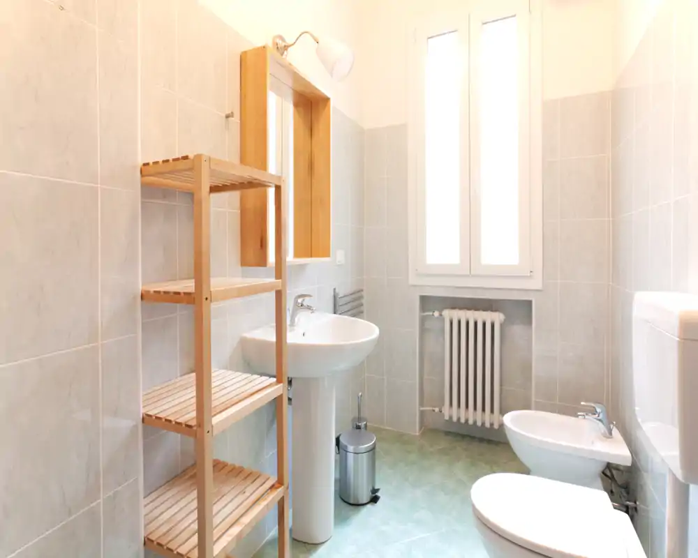 Baño luminoso y bien iluminado con lavabo de pedestal, bidé, inodoro, radiador toallero y estantería de madera; suelo verde menta y una amplia ventana translúcida que aporta luz natural.