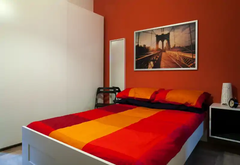 Dormitorio luminoso y acogedor con cama doble con ropa de cama roja y naranja, mesita y cuadro en la pared — limpio y agradable.