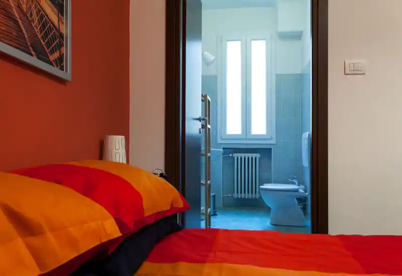 Rincón de dormitorio luminoso y acogedor con una cama colorida en primer plano y baño en suite visible a través de la puerta abierta.