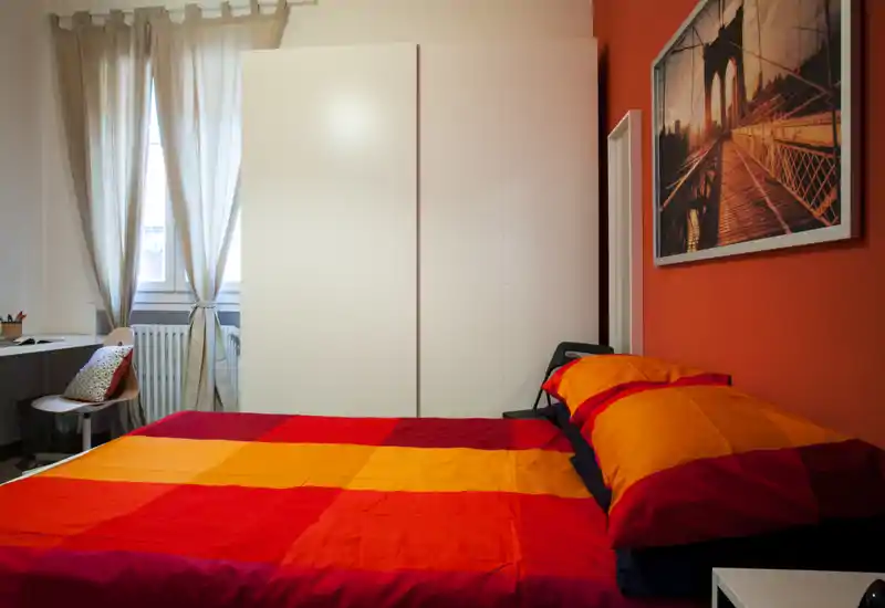 Dormitorio luminoso y acogedor con una cama doble grande con ropa de cama roja y naranja, armario blanco, escritorio junto a la ventana y cuadro decorativo. La luz natural que entra por las cortinas translúcidas aporta una sensación agradable.