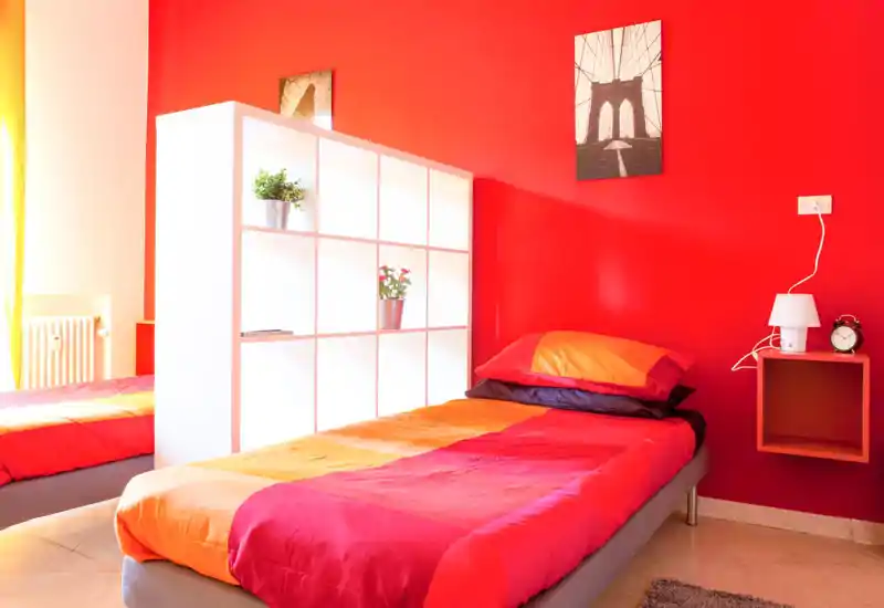 Dormitorio individual moderno y luminoso con edredón rojo-naranja, balda de noche con lámpara y despertador, y estantería blanca modular con macetas — ideal para un alquiler juvenil y alegre.