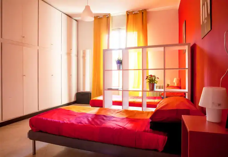 Dormitorio luminoso y acogedor en tonos rojos y naranjas, cama visible en primer plano, armarios empotrados y ventana con cortinas amarillas que proporcionan mucha luz natural.