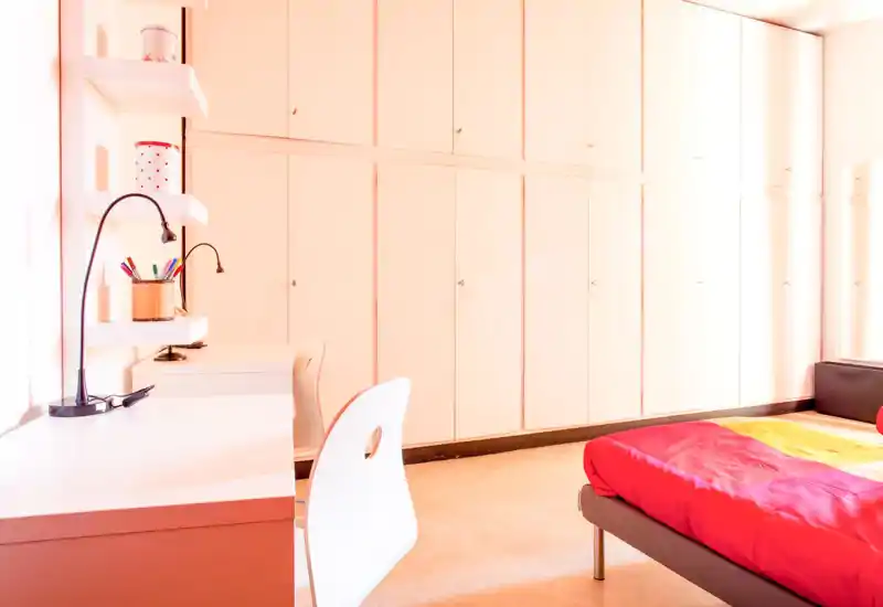 Dormitorio moderno y luminoso con cama baja y ropa de cama roja intensa, zona de estudio con estantes y lámparas de escritorio, y una pared de armarios empotrados que ofrecen amplio almacenamiento.