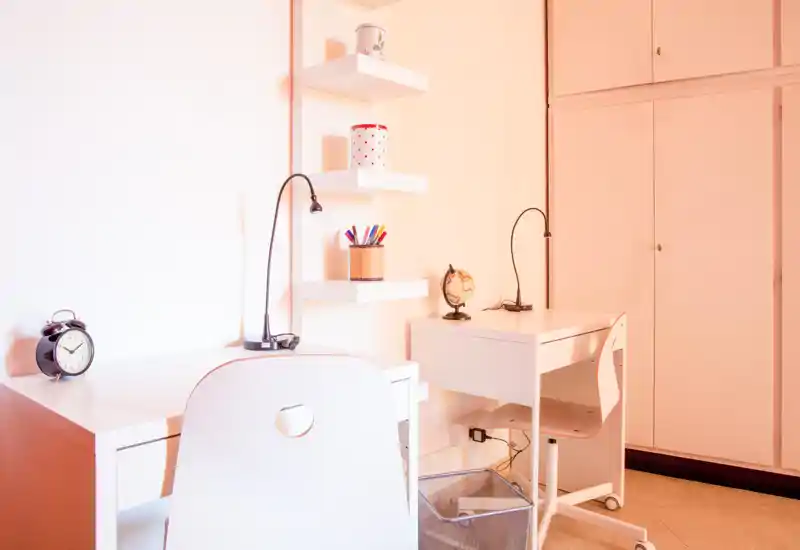 Rincón de estudio luminoso y minimalista con dos escritorios blancos, lámparas de lectura y estantes — un espacio de trabajo tranquilo en un ambiente tipo dormitorio.