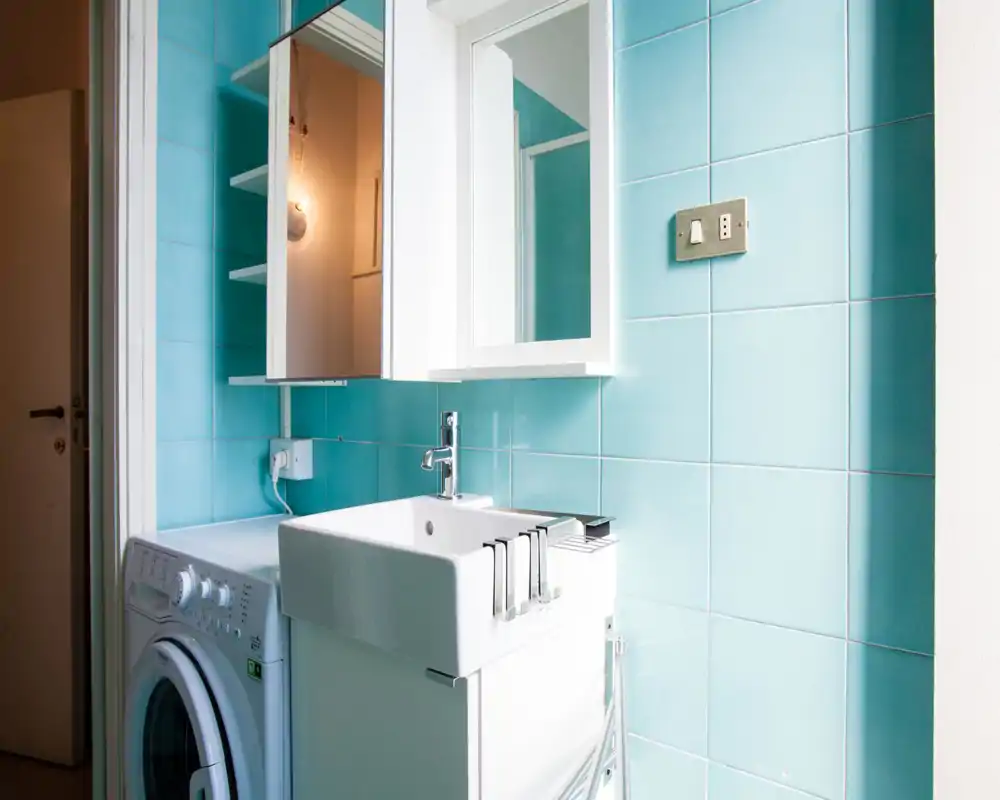 Bagno piccolo e luminoso con piastrelle turchesi, lavabo bianco e specchiera; una lavatrice è posizionata accanto al mobile del lavabo.