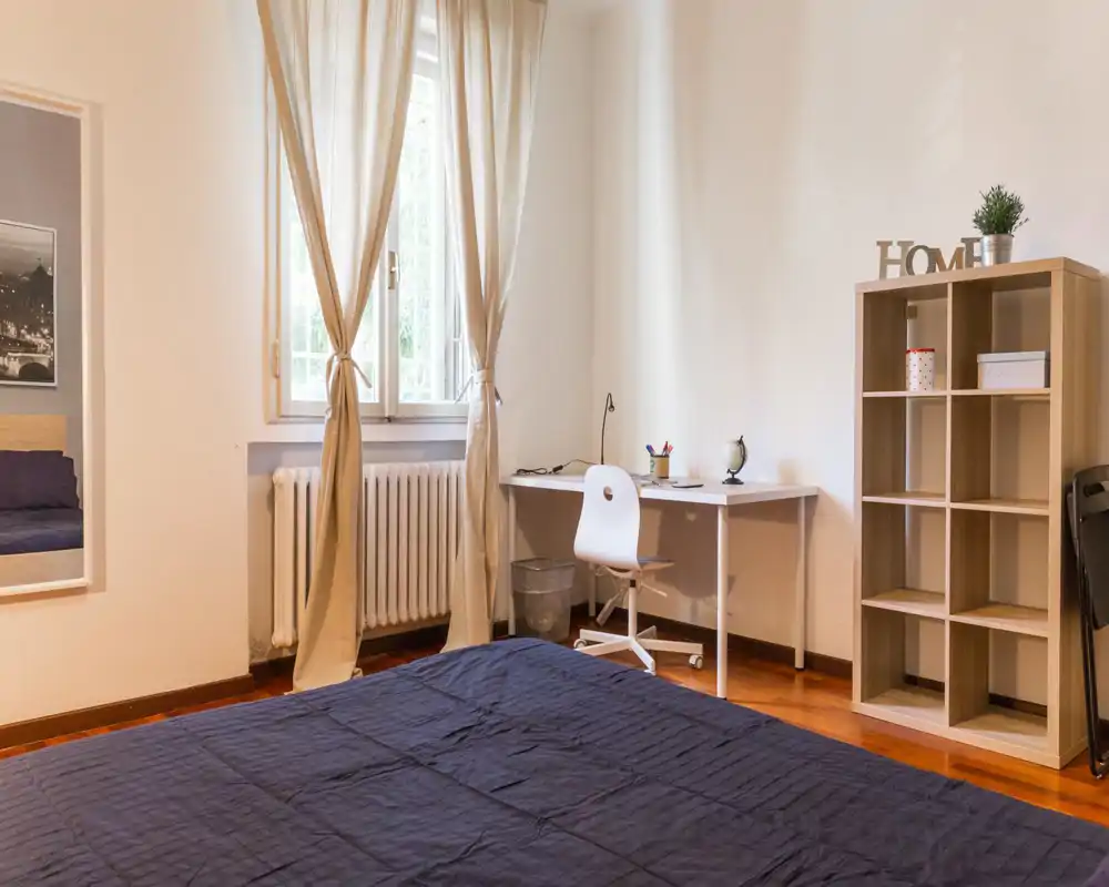 Chambre lumineuse et soignée avec lit double au premier plan, bureau près de la fenêtre et étagère à cases — idéale pour un étudiant ou un professionnel recherchant une pièce simple et bien éclairée.