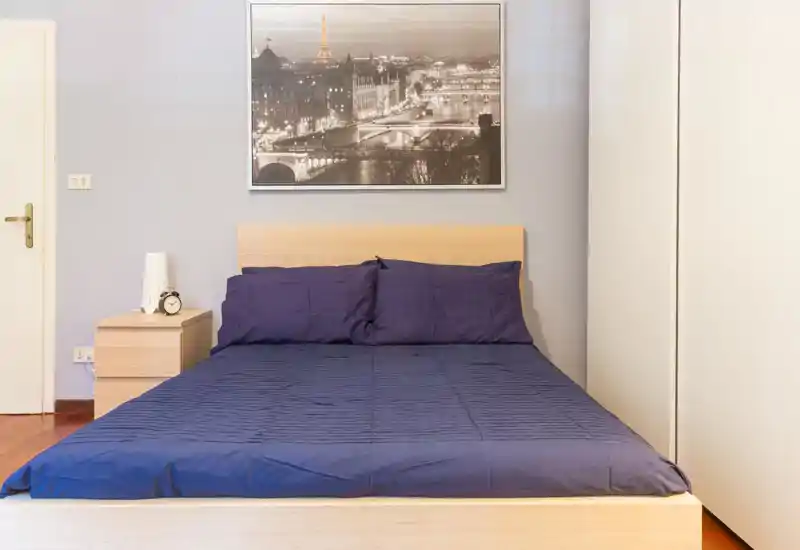 Chambre lumineuse et bien rangée avec un lit double, tête de lit en bois et table de chevet assortie ; décoration moderne et sobre avec une affiche de ville au-dessus du lit.