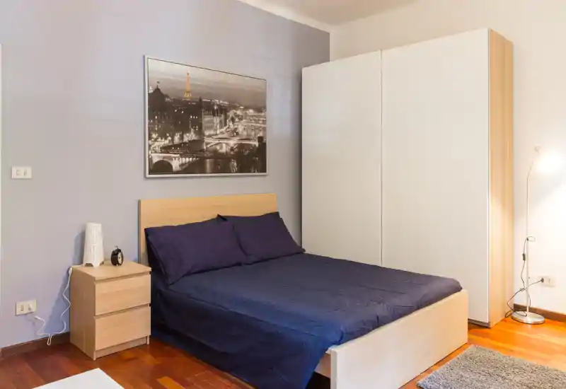 Chambre lumineuse et bien mise en scène avec lit double, table de chevet et armoire ; linge de lit bleu marine et sol en bois pour une ambiance chaleureuse et moderne.