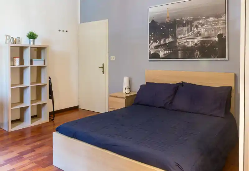 Chambre moderne et lumineuse avec lit en bois et linge de lit bleu marine, table de chevet avec réveil ; une étagère et une grande photo encadrée apportent style et praticité.