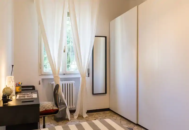 Angolo camera da letto luminoso e ordinato con letto a righe in primo piano, grande armadio bianco, scrivania con lampada e sedia vicino alla finestra con tende — toni neutri e luce naturale.