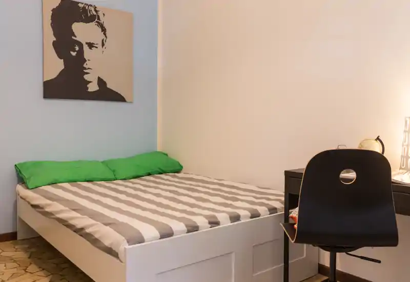 Habitación compacta y ordenada con cama doble con ropa de cama a rayas, almohadas verdes y un pequeño escritorio con silla al lado — luminosa y de estilo sencillo.