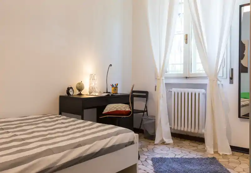 Dormitorio luminoso y ordenado con cama doble con ropa de cama a rayas, un pequeño escritorio con lámpara y globo, y una ventana con cortinas claras que aporta luz natural.