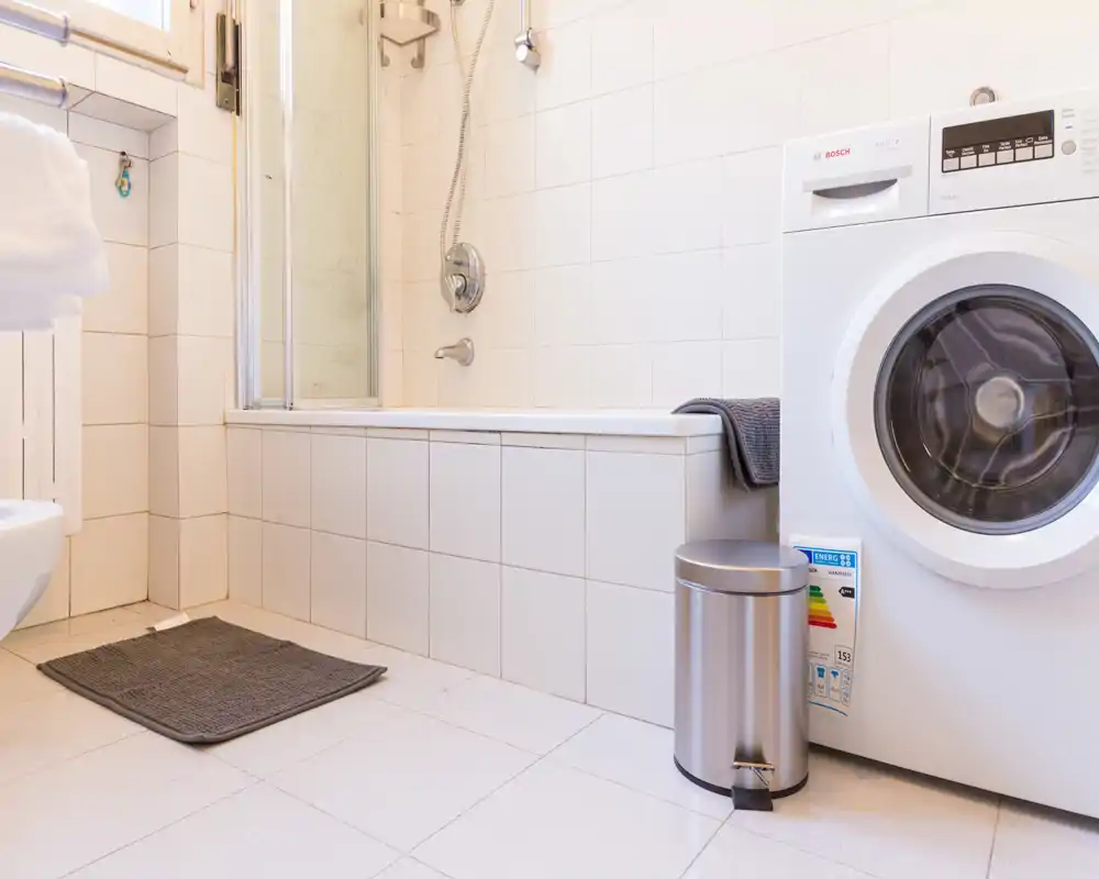 Baño limpio y luminoso con bañera y ducha integrada; lavadora y un pequeño cubo de acero inoxidable completan la disposición funcional.
