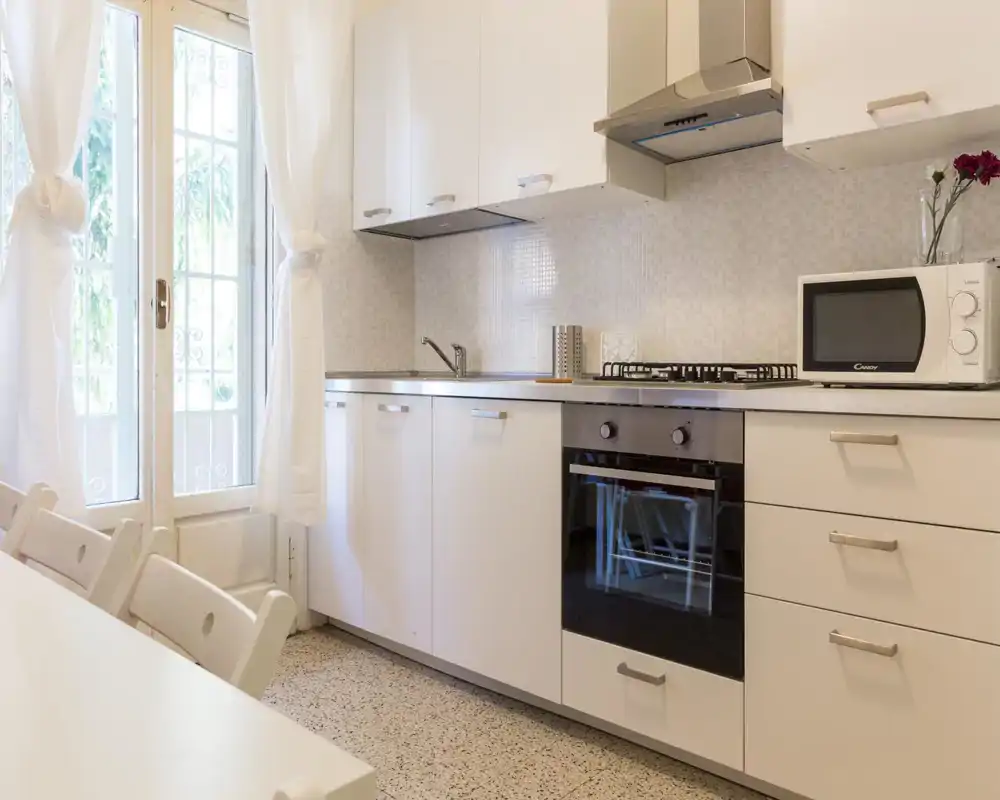 Cocina moderna y luminosa con muebles blancos, horno empotrado y placa de gas, microondas y fregadero; sensación de amplitud por la puerta acristalada y las cortinas claras.
