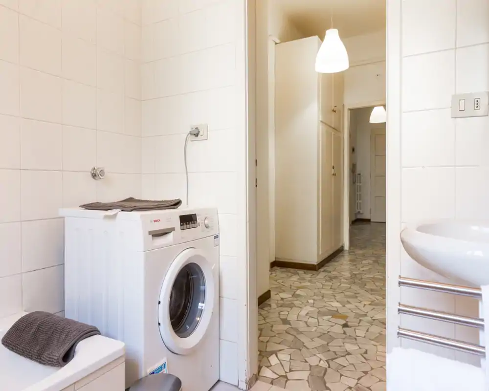 Baño limpio y luminoso con lavadora, lavabo y paredes alicatadas; pasillo visible a través de la puerta.