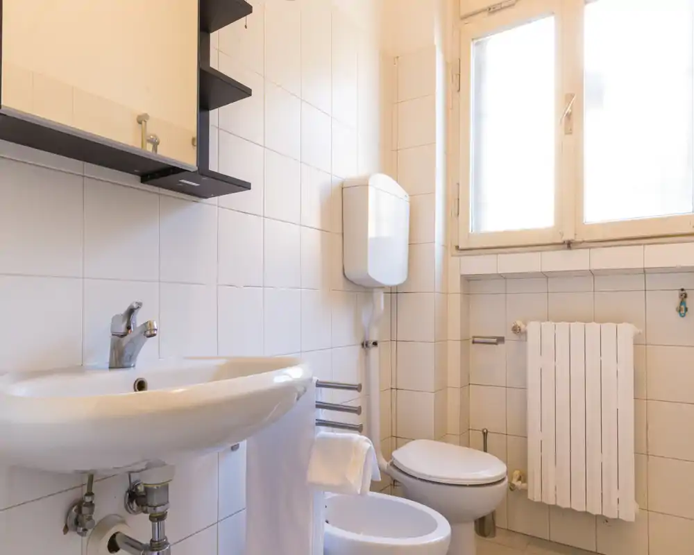 Baño compacto y bien iluminado con lavabo, bidé, inodoro y una gran ventana esmerilada que aporta luz natural. Azulejos blancos y un armario de pared ofrecen almacenamiento práctico.