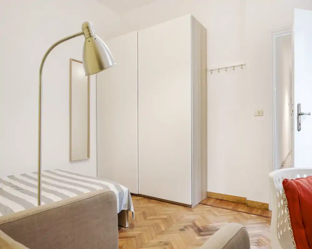 Chambre lumineuse et bien rangée avec lit double, lampadaire et grand placard blanc. Le parquet chaleureux et les textiles apportent une atmosphère accueillante, idéale pour une annonce.