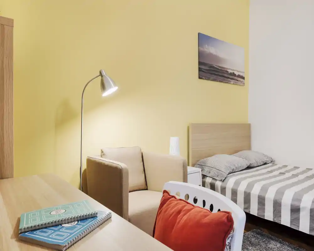 Chambre simple lumineuse et bien aménagée avec lit à rayures, fauteuil confortable et bureau — idéale pour étudiant ou voyageur seul.