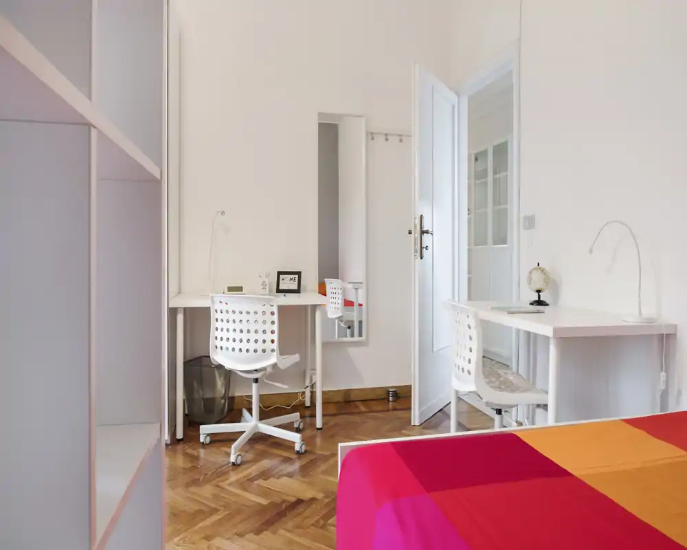 Bright, tidy bedroom with a colorful bedspread, two white desks and chairs, and a mirror on the wall. Hardwood parquet and minimal modern décor create a fresh, airy feel ideal for a student or guest room.