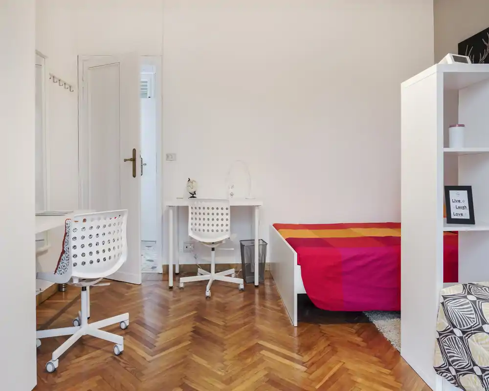 Bright, neatly styled bedroom with a single bed featuring colorful bedding, a study desk and chair, and clean parquet flooring — ideal for a student or single occupant.