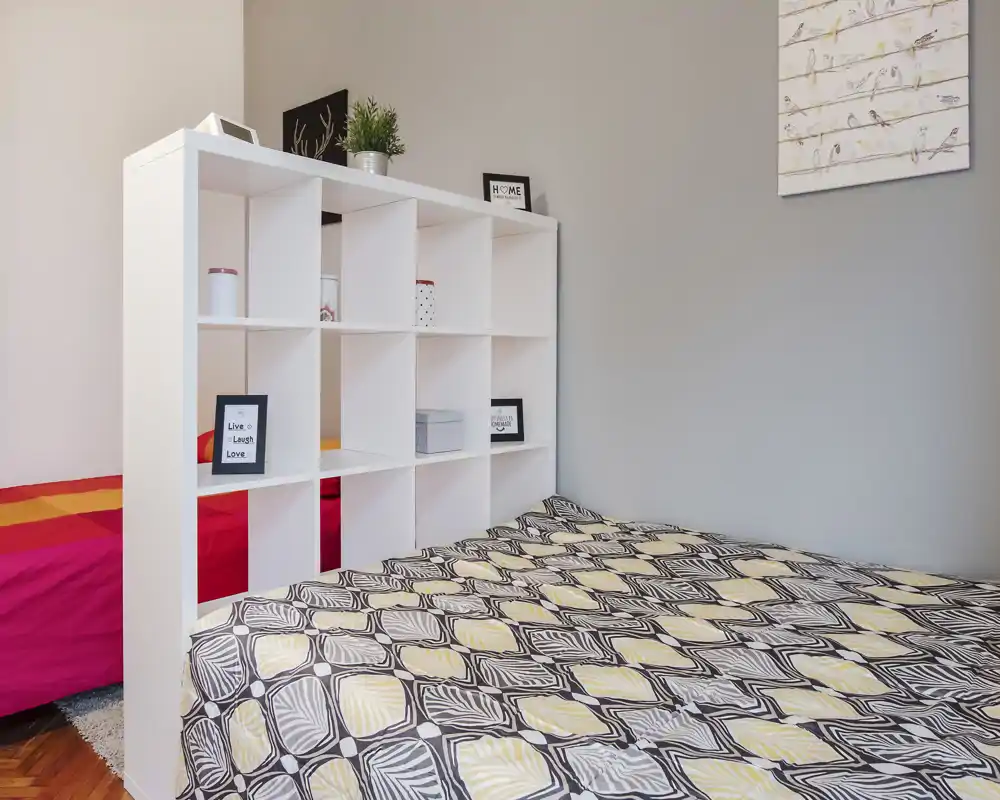 Comfortable bedroom with a large patterned bed in the foreground and a white open-shelf room divider. Neutral walls and soft lighting create a calm, inviting atmosphere.