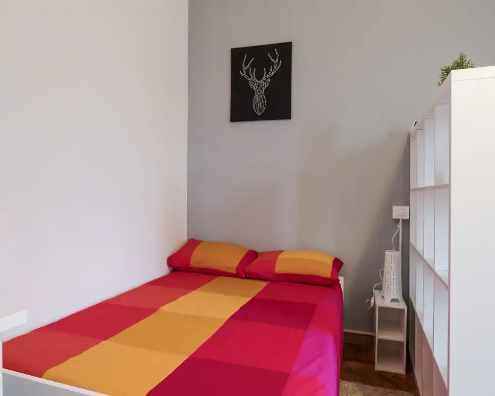 Bright, neatly styled bedroom with a double bed dressed in warm red and orange bedding, a small shelving unit and minimalist wall art — ideal for a cosy short-term stay.