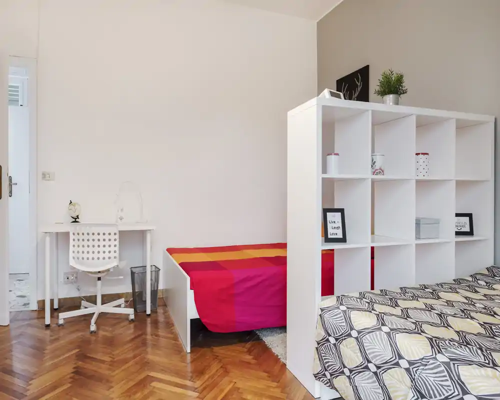 Bright, neatly staged bedroom with two beds separated by a white shelving unit; patterned duvet in the foreground and a colorful single bed in the background create a warm, modern feel.