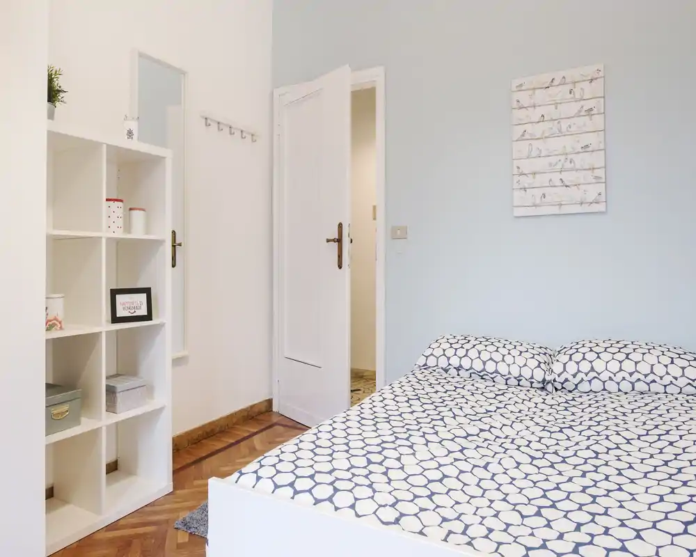 Dormitorio luminoso y ordenado con ropa de cama estampada, una estantería blanca y pared en tono pastel; suelo de parquet y cuadro decorativo que aportan tranquilidad.