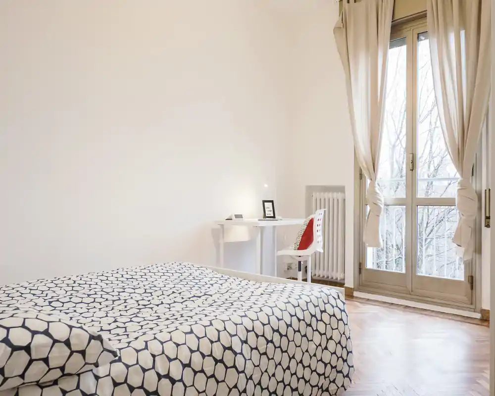 Dormitorio luminoso y minimalista con ropa de cama estampada, un pequeño escritorio junto a la ventana y abundante luz natural de las puertas balconeras.