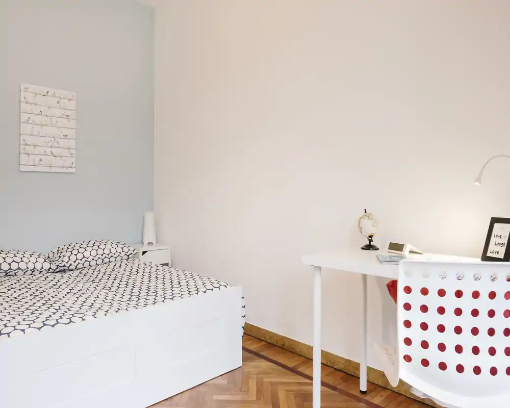 Dormitorio luminoso y minimalista con cama blanca y ropa de cama estampada, además de un escritorio y silla sencillos — espacio acogedor y bien iluminado, ideal para una persona o estancias cortas.