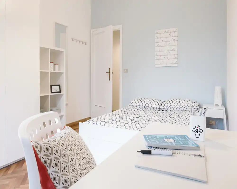 Dormitorio luminoso y minimalista con una cama doble con ropa de cama estampada, mesita con lámpara y un escritorio ordenado en primer plano — ideal para una estancia tranquila.