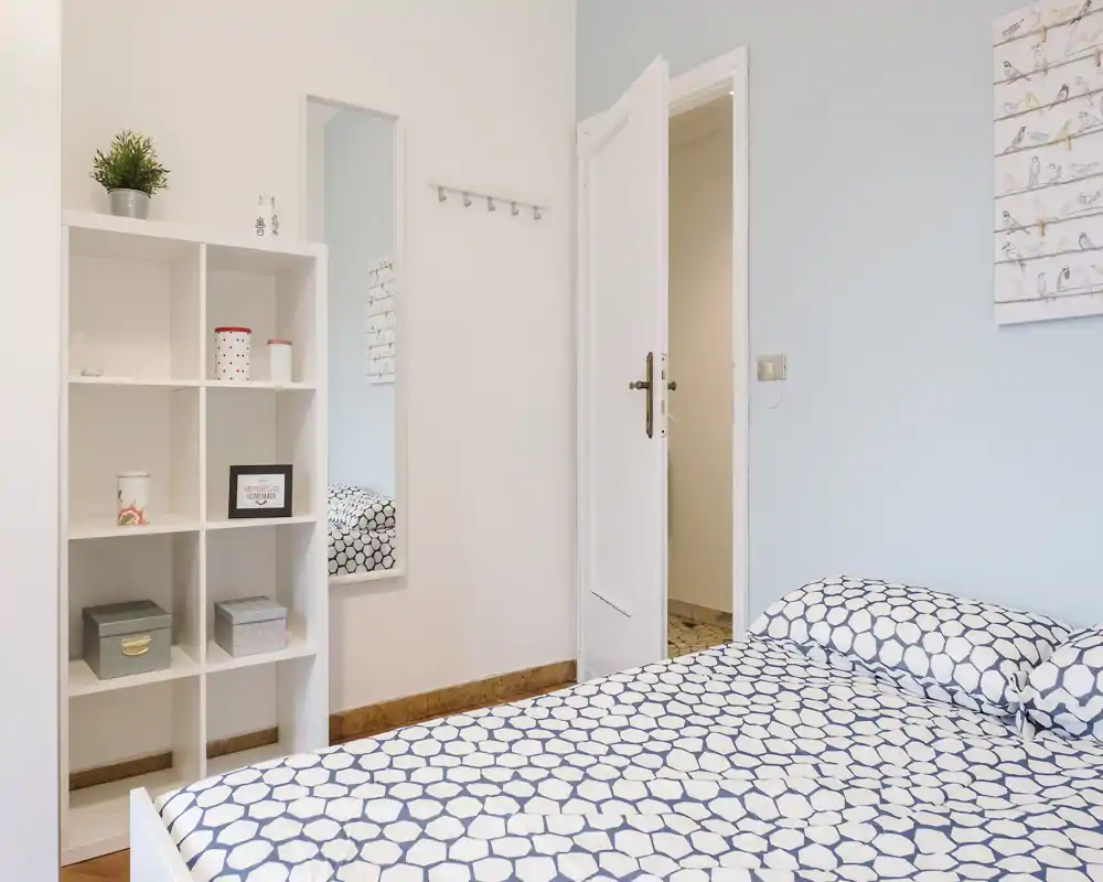 Dormitorio luminoso y ordenado con ropa de cama estampada, una estantería blanca y un espejo de cuerpo entero — decoración limpia y minimalista ideal para alquileres.