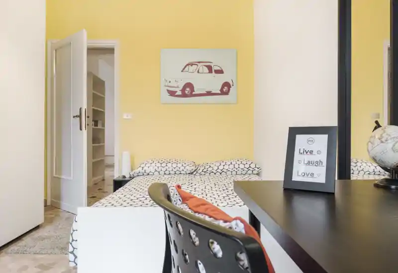 Camera da letto luminosa e moderna con biancheria a fantasia, quadro decorativo e una piccola postazione di lavoro in primo piano. L'ambiente è accogliente e ben illuminato, ideale per affitti brevi.