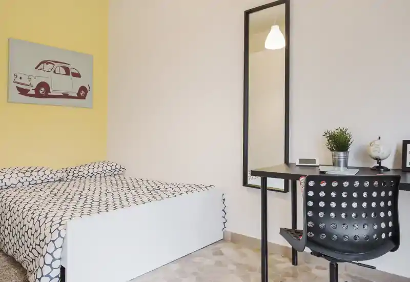 Camera da letto luminosa e moderna con letto dalla struttura bianca, biancheria a motivo e una piccola scrivania con specchio — ambiente ordinato e ben illuminato, ideale per gli ospiti.
