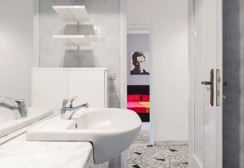 Bagno moderno e luminoso con lavabo bianco, rubinetteria cromata e mobile con specchio; dalla porta si intravede una camera dai colori vivaci.