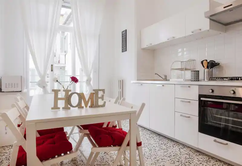 Cucina moderna e luminosa con mobili bianchi, forno a gas e tavolo da pranzo con cuscini rossi e lettere decorative "HOME", illuminata da luce naturale attraverso ampie finestre.