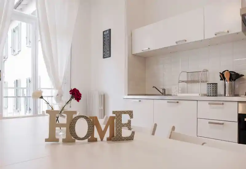 Cucina open-space luminosa e moderna con mobili bianchi, piano di lavoro pulito e tavolo decorato con fiori e lettere decorative 'HOME'.