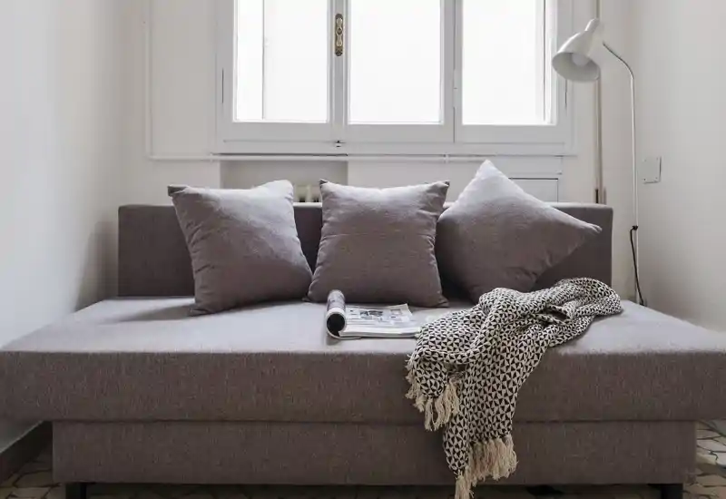Angolo sitting luminoso e minimale con divano-letto compatto, tre cuscini e un plaid decorativo sotto una finestra dalla luce morbida — un angolo accogliente per rilassarsi o leggere.
