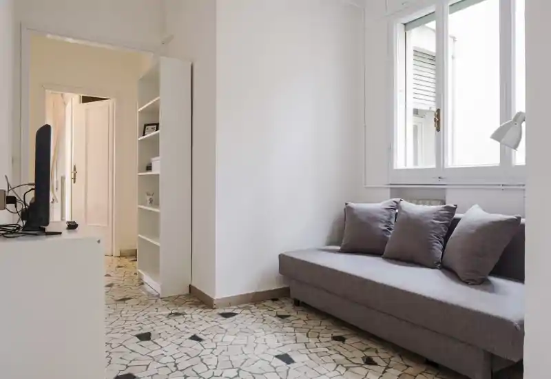 Soggiorno compatto e luminoso con divano grigio moderno, cuscini e luce naturale da una grande finestra — ideale per un soggiorno cittadino accogliente.