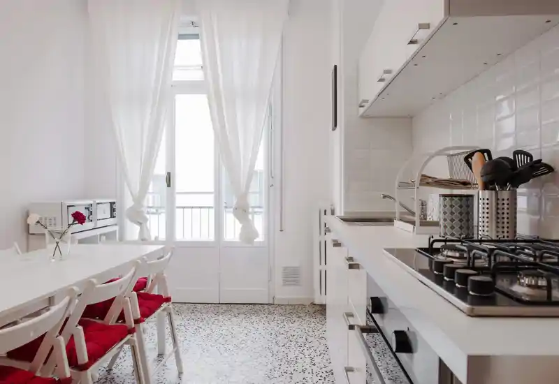 Cucina moderna e luminosa con pensili bianchi, piano cottura a gas e utensili sul piano; tavolo da pranzo con sedie con cuscini rossi accanto a una grande finestra che illumina l'ambiente.