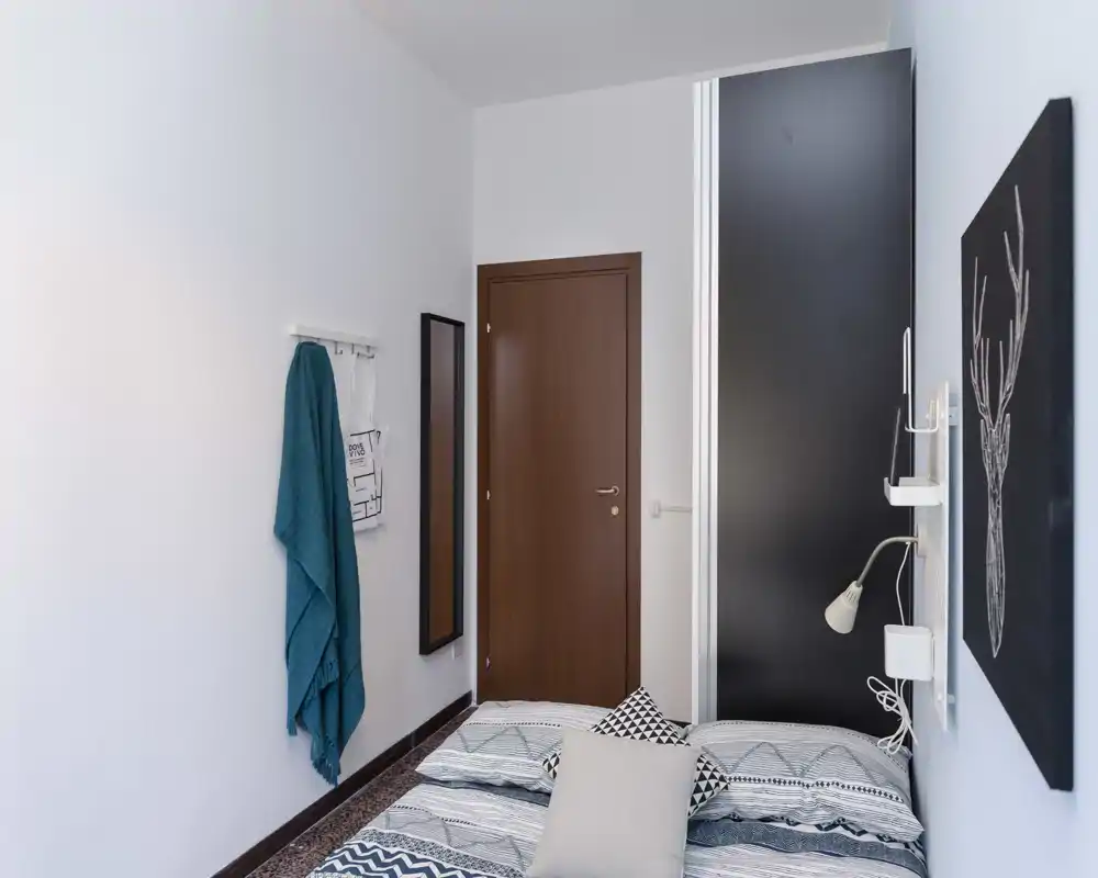 Chambre compacte et lumineuse avec couette à motif, coussins et décoration murale sobre ; placard et porte encadrent le lit, adaptée pour un séjour cosy.
