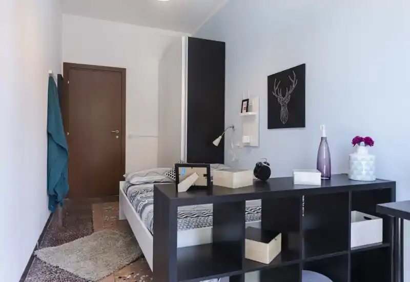 Chambre lumineuse et bien décorée avec un lit double, une lampe de chevet et des étagères décoratives ; meuble de rangement noir au pied du lit apportant séparation et espace d'exposition.